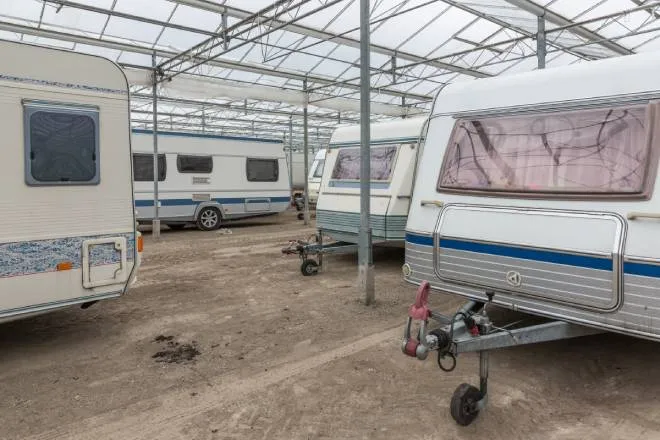 An RV storage facility contains numerous caravans parked on a dirt lot. They are covered by a large tarp acting as a roof.