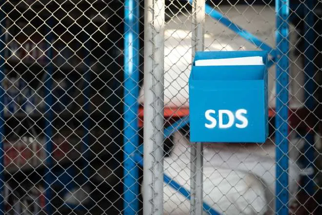 A blue metal box labeled "SDS" hangs on a chain-link fence and holds documents. Storage containers are behind the fence.