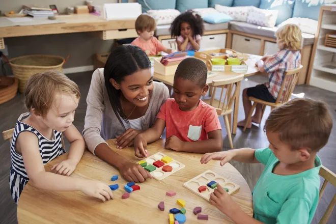 Young children and a care giver at tables engaged in educational activities.