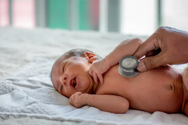 Newborn baby on a bed with hand holding a stethoscope to the baby's chest.