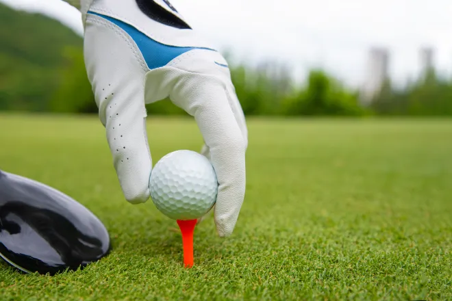 Close up of a gloved hand setting a golf ball on a tee. A golf club is near the tee.