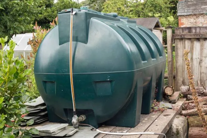 A green water tank is in the middle of a garden on a wooden platform. Behind it is a fence.