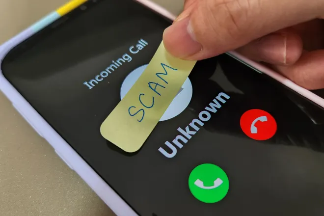 Smartphone showing an incoming call from an unknown number. Fingers are placing a hand-written label with the word "scam" over the top.