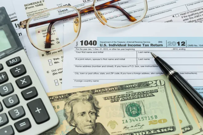 Closeup of United States currency, calculator, pen, and glasses on top of tax forms.