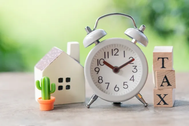 Analog alarm clock flanked by a stack of wooden blocks with the word "tax" and a small house figurine.