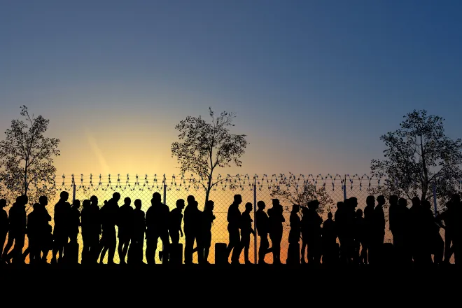 Silhouetted line of people at a fence topped with razor wire at sunrise or sunset.