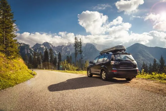 A car with a hard shell roof cargo carrier parked on the side of a winding mountain road with clouds in the sky.