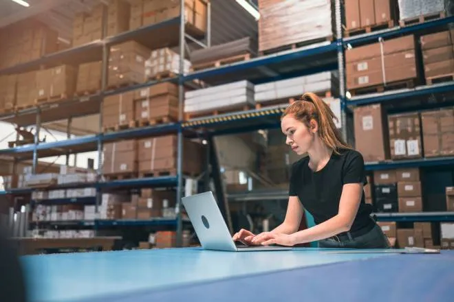 A woman with a ponytail wearing a black shirt standing in front of a table using a laptop in a warehouse.