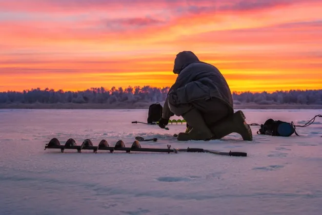 A man kneeling on an icy snow-filled field preparing an ice fishing hole with an auger drill attachment on the ground.