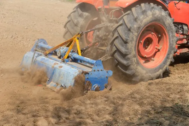 A large red tractor with a blue rotary tiller attachment The tiller is kicking up clouds of dirt in a field.