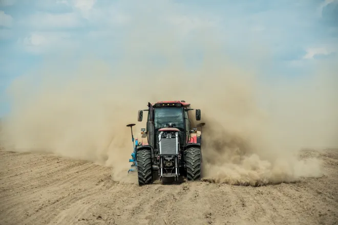 Farm tractor driving through bare dirt creating a dust cloud.