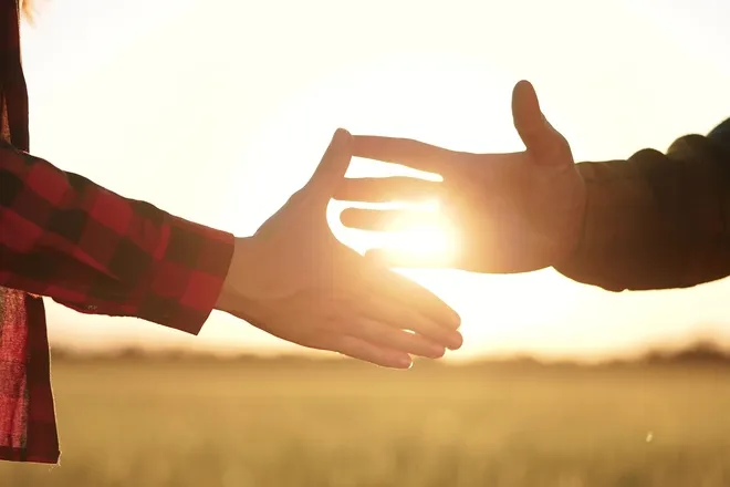 Two people shaking hands as the sun goes down behind them. They're standing on a piece of empty farmland.