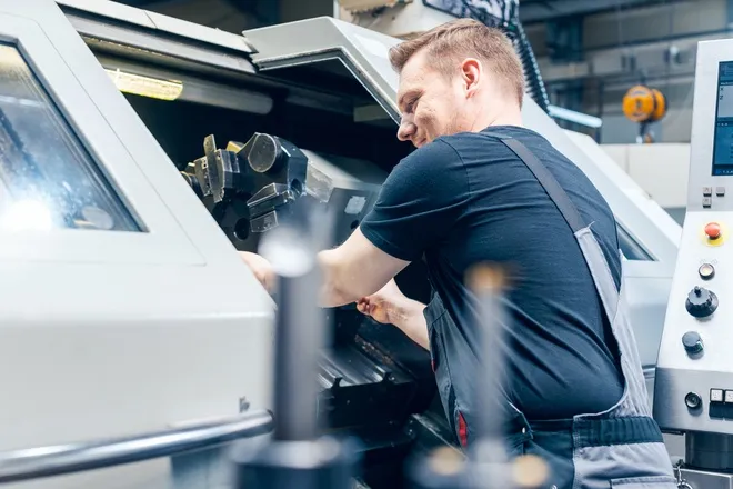 A professional tooling machine worker in a warehouse. They're smiling as they work on a tooling project inside the machine.