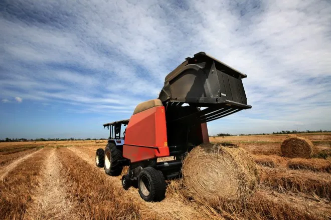 A rear view of a blue tractor towing a red round hay baler and dropping a rolled bale of hay onto a harvested field.
