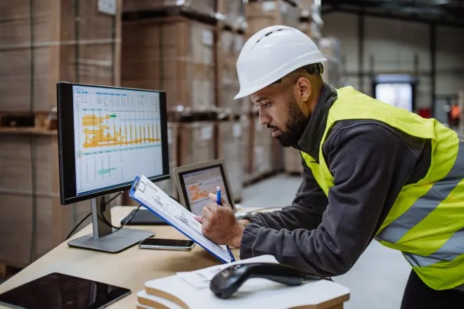 A warehouseman, wearing a neon yellow vest and a white hard hat, is checking the delivery and stock on a computer.