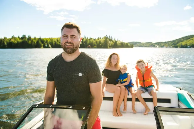 A man stands behind the steering wheel of a boat. A woman and two children sit behind the man at the back of the boat.