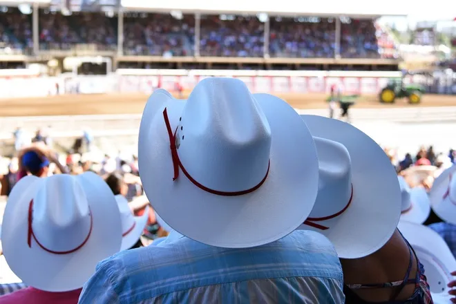 Three people in matching white cowboy hats adorned with red ribbons sitting among a crowd in the stands at a rodeo.
