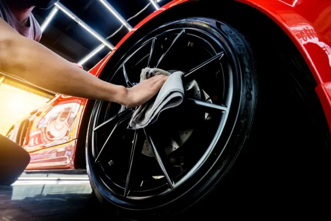 A man is using a washcloth to clean the spokes of a car tire. The car is red and the man’s face is obscured by a mask.