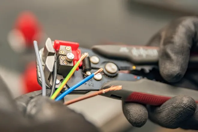 A close-up of a pair of hands with black gloves using a manual wire stripper to strip five colored electrical wires.