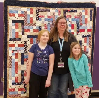 Woman and two children standing in front of a quilt.