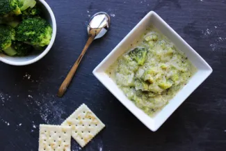 Square bowl of Cream of Broccoli Soup, a bowl of broccoli, two crackers and a spoon.