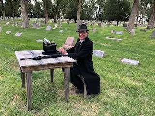 Tim Baublits portraying George Wick, editor of the Bent County Democrat from 1899 to 1930. Man seated at a table in cemetery with a manual typewriter.