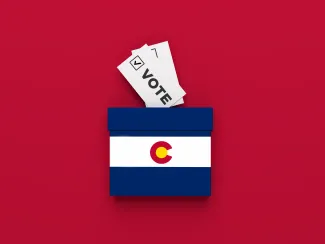 Miniature ballot box with and image of the Colorado state flag on a red background with slips of paper representing ballots