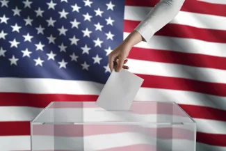PROMO HIRES Miscellaneous - Election Vote Ballot Flag - iStock - sefa ozel Hand placing a piece of pager into a clear ballot box in front of the United States flag