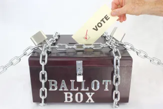 Hand inserting a piece of paper labeled "vote" into a wood box labeled "Ballot Box." The box is secured by chains