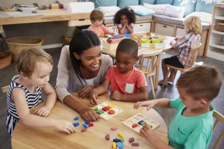 Young children and a care giver at tables engaged in educational activities.