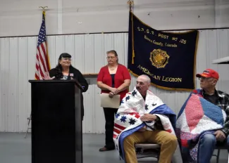 Ceremony to present quilts to veterans. Two speakers are standing, and two recipients are seated.