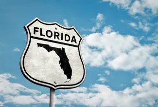 Road sign with the world "Florida" and a silhouette of the state against a clouded sky background.