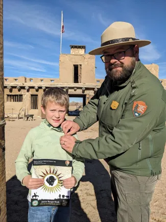 A Park Ranger pins a badge on a young Junior Ranger, who is holding a copy of the park's new Junior Ranger Book. A Park Ranger pins a badge on a young Junior Ranger, who is holding a copy of the park's new Junior Ranger Book.