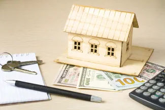 PROMO HIRES Finance - Money House Calculator Keys Pen Paper - iStock - Motizova Model house made of wood resting on United State currency. A calculator, pen, notepad, and keys are nearby.