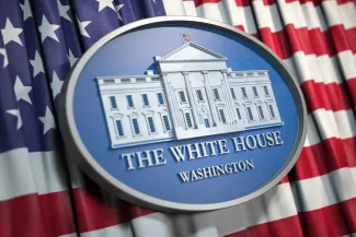 PROMO HIRES CROPPED Government - Politics White House Washington DC Flag United States - Bet_Noire - iStock-1264844457 Close up of an oval, carved plaque with an image of the White House. The plaque is on top of a flag of the United States of America.
