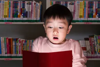 Child with a surprised expression holding an open book with glowing pages in front of books on shelves