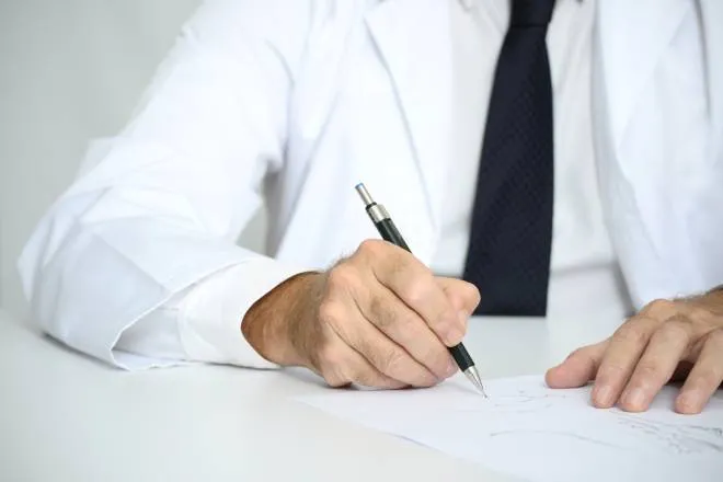man in a medical coat, button down shirt, and tie writing on a piece of paper with a black mechanical pencil