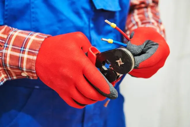An electrician wearing red working gloves crimping multiple electrical wires with a hand crimping tool.