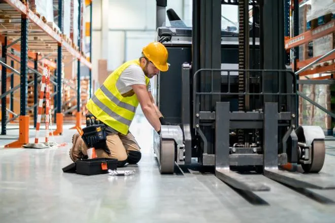 A mechanic kneeling next to a forklift in a warehouse as he makes repairs with a tool kit by his side.