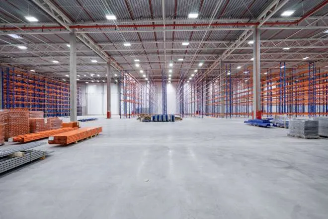 A large empty warehouse with high beam, LED lights. There are empty blue and orange rafters for inventory.