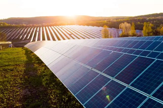 A row of solar panels on a solar farm perched on a hill. The sun is setting in the background, casting a bright glow.