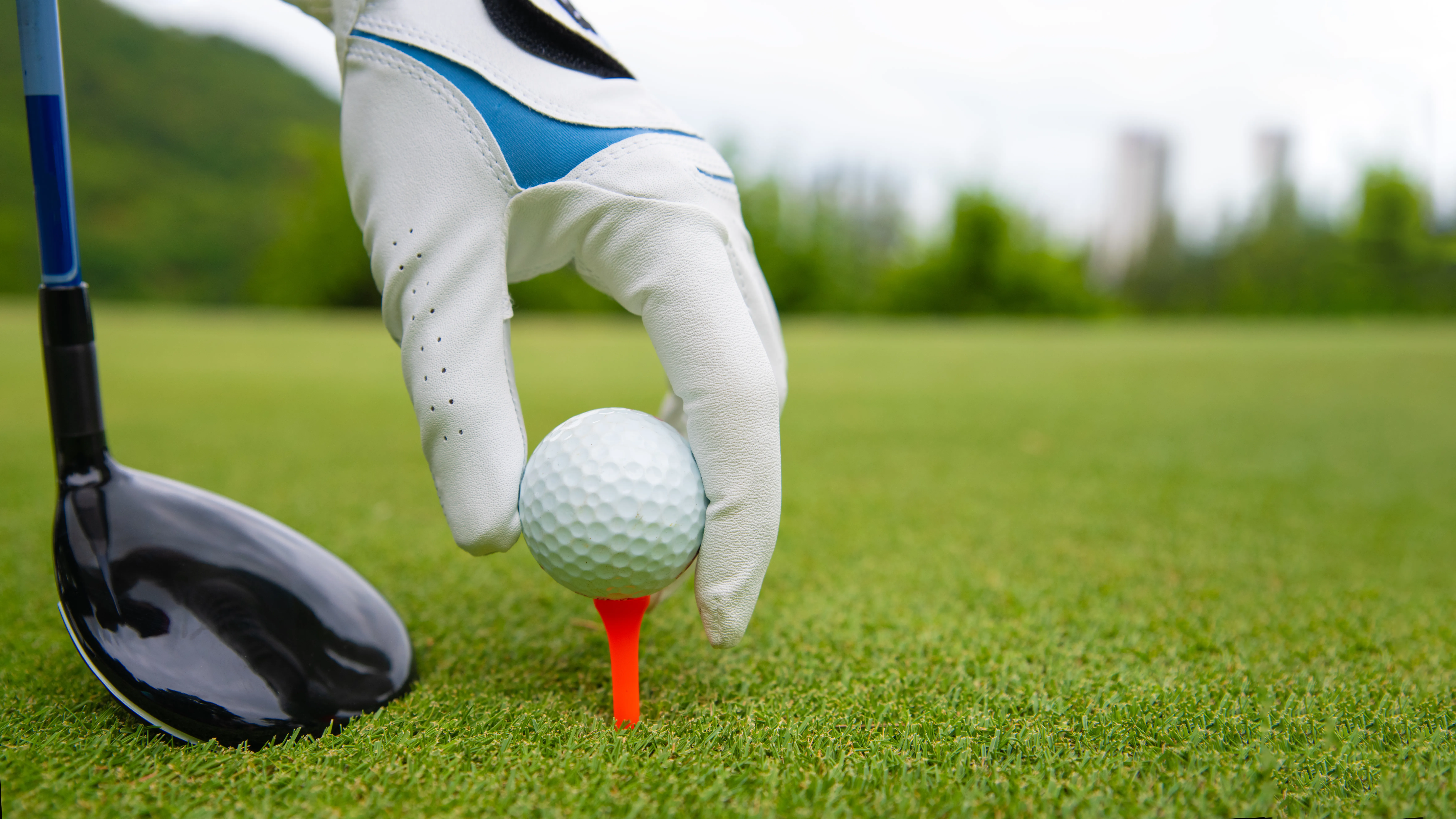 Close up of a gloved hand setting a golf ball on a tee. A golf club is near the tee.