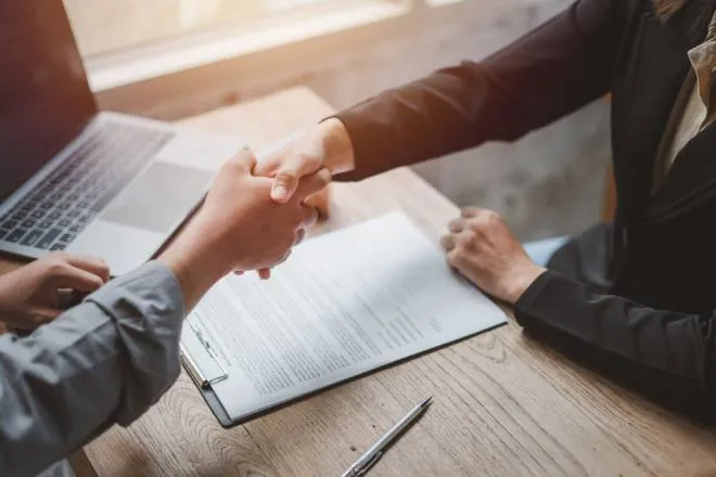 A buyer and a seller sitting at a table together shaking hands after reaching a deal. The contract is sitting on the table.