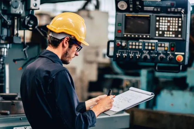 A team member wearing a blue jumpsuit and a yellow hard hat stands next to an automation machine, writing on a notepad.