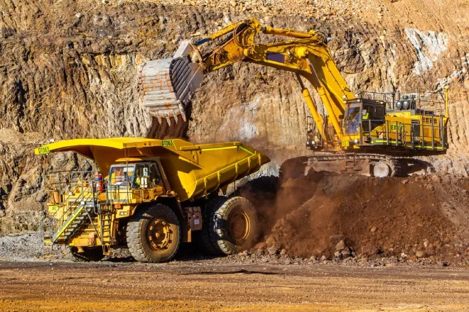 Two mining vehicles work together at a mining site. A digger on top of a small hill loads a dump truck with material.