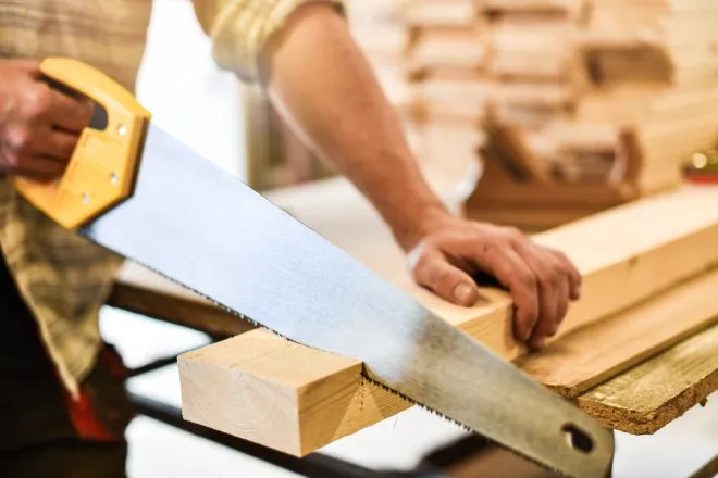 A close-up of a man using a large hand saw to cut a long piece of wood. He is holding the wood in place while he saws.