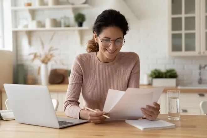 A female homeowner sitting at the dining table reading through printed tax documents. She has a laptop in front of her.