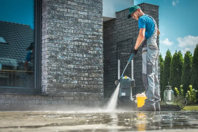 A man wearing overalls uses a pressure washer to clean the concrete patio outside of a large brick home.