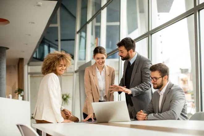 Four employees in business attire are clustered together around a laptop, looking at the screen.