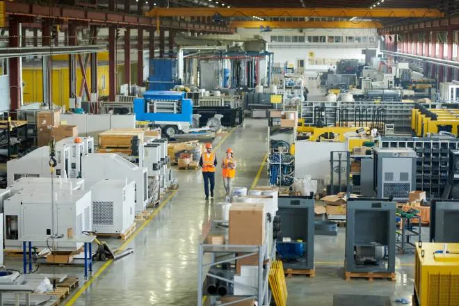 A large factory of machinery and heavy equipment with two workers in orange high-visibility vests and helmets walking through.